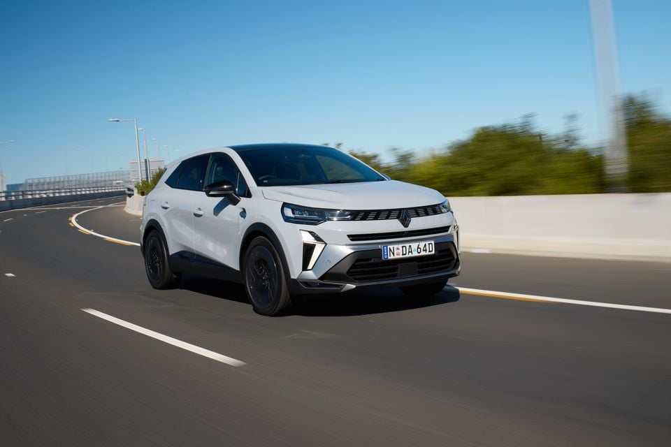A white 2026 Renault Symbioz SUV driving on a road, seen from the front-side