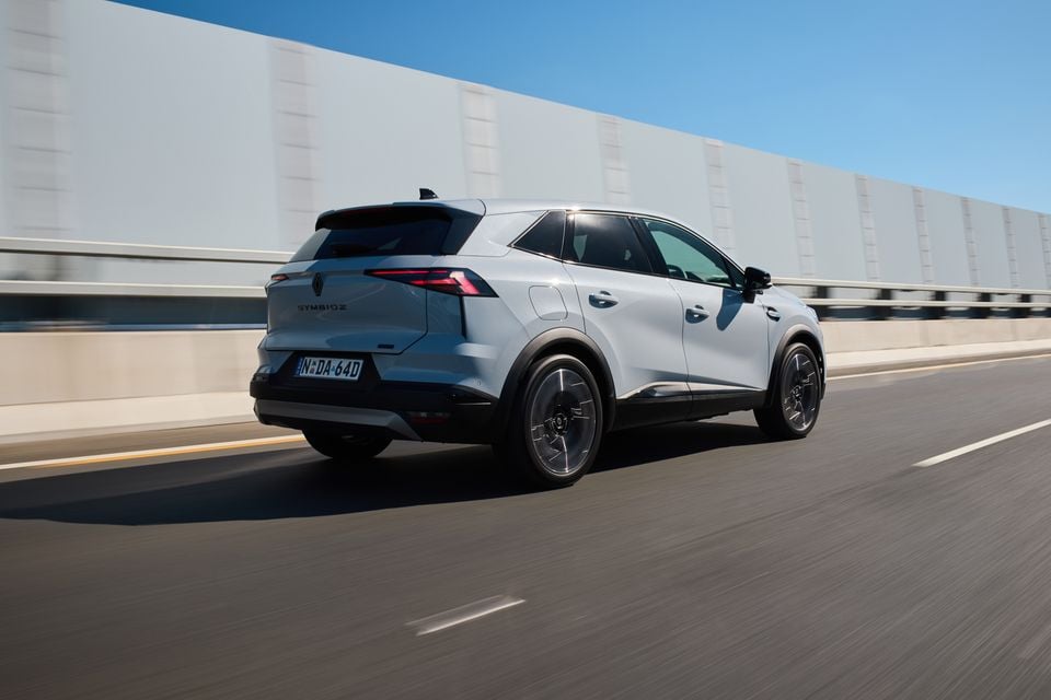 A light blue 2026 Renault Symbioz SUV seen from the rear three-quarter angle while driving on a highway