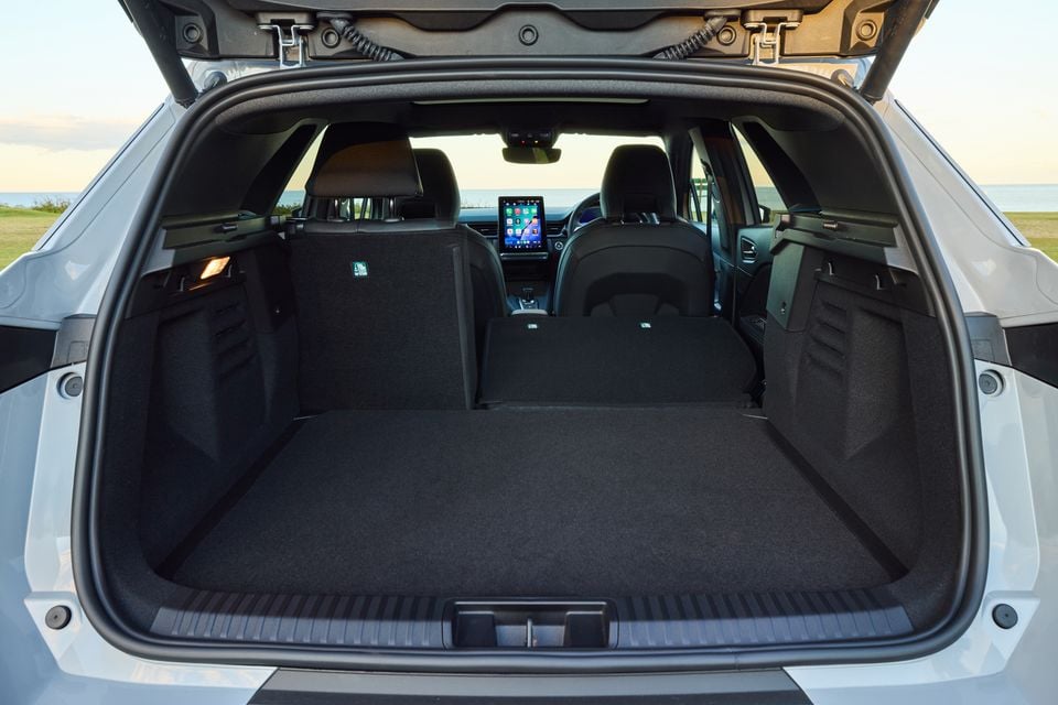 The open boot of a 2026 Renault Symbioz SUV showing a large, empty cargo area with the rear seats folded, viewed from the rear with a coastal backdrop.
