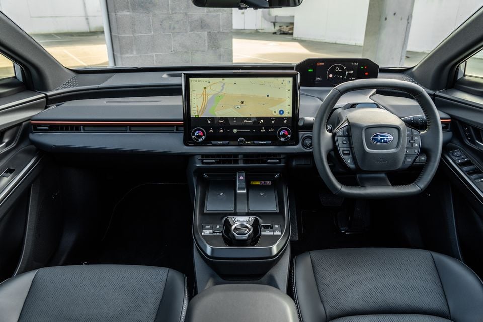 Subaru Solterra interior showing the dashboard, large central touchscreen displaying navigation, digital instrument cluster, flat-bottom steering wheel with Subaru badge, and centre console with wireless charging pads