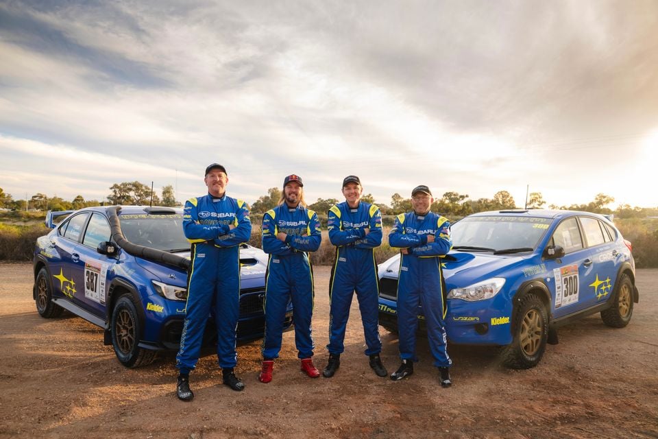 Four rally drivers in Subaru racing suits pose with two blue Subaru rally cars numbered 387 and 300 at the Sunraysia Safari Rally