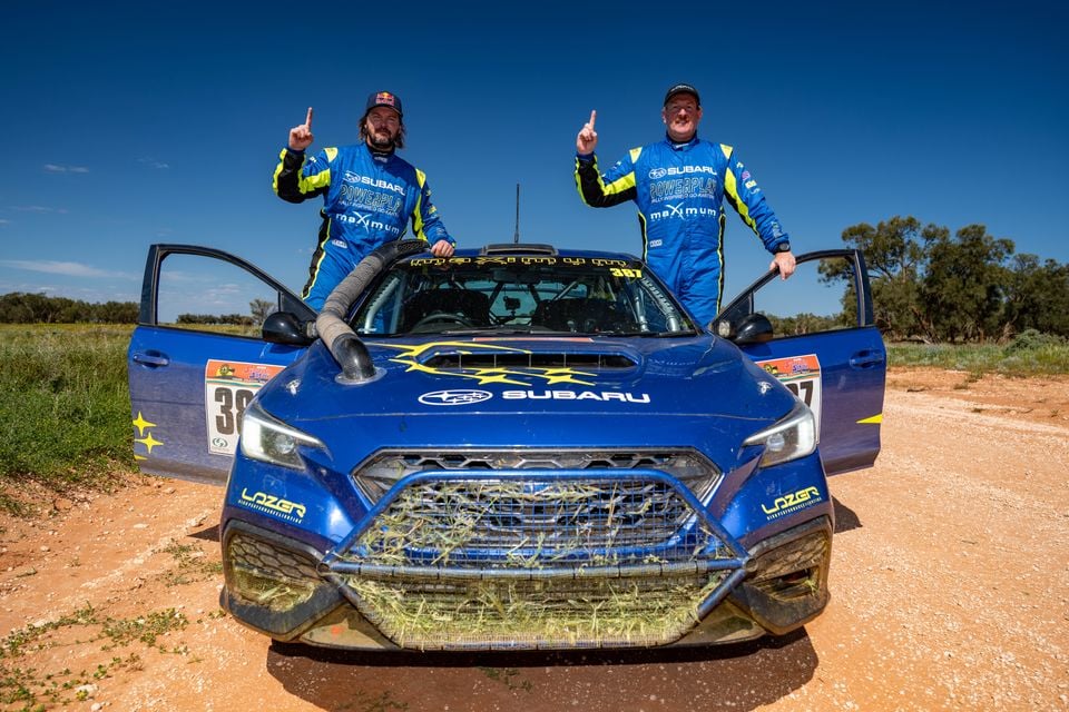 Two rally drivers in Subaru Powerplay racing suits celebrate with a number-one gesture on the bonnet of a mud-covered blue Subaru WRX rally car, number 387, on a dirt track in outback Australia