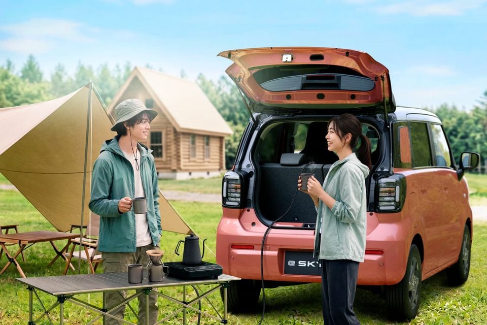A pink Suzuki e:Sky kei car with its tailgate open at a campsite, with two people holding mugs and a portable coffee setup on a folding table nearby