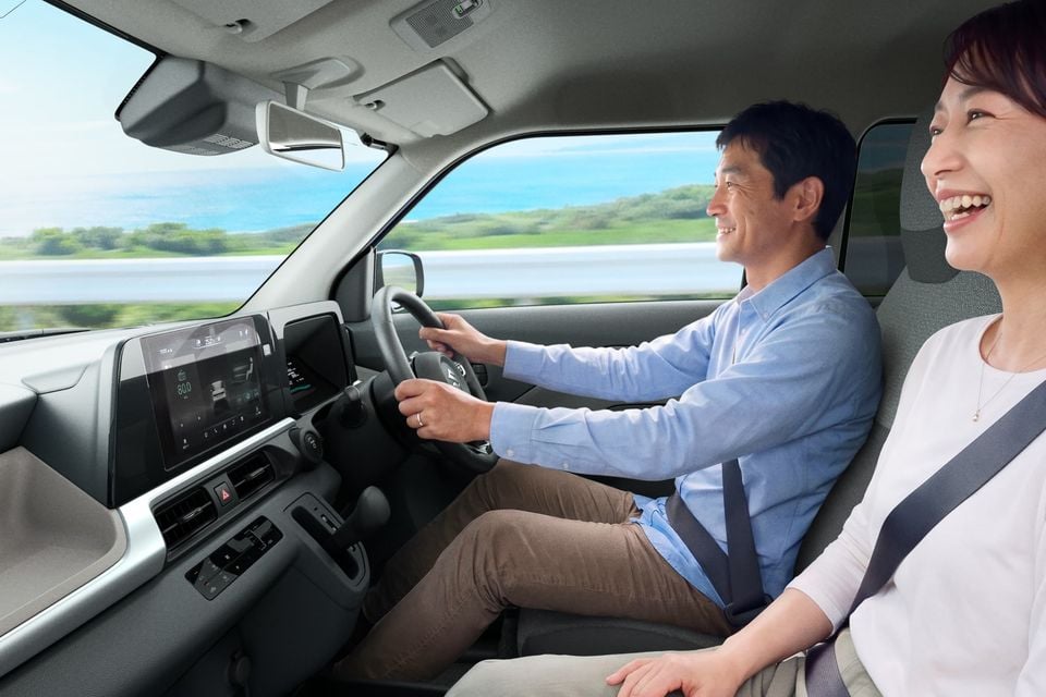 Two passengers smiling inside a Suzuki e:Sky, with the driver at the wheel and a touchscreen infotainment display visible on the dashboard