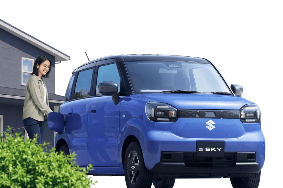 A blue Suzuki e Sky compact electric car seen from the front three-quarter angle, with a woman standing beside it outside a house