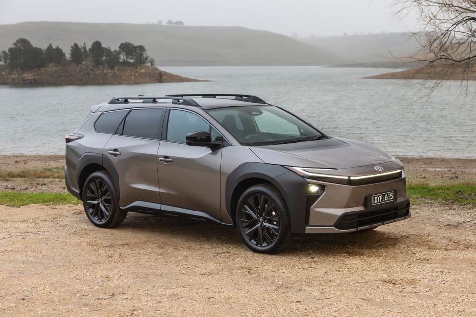 A grey 2026 Toyota bZ4X PHEV Touring SUV seen from the front-side, parked on a gravel area beside a lake on a misty day