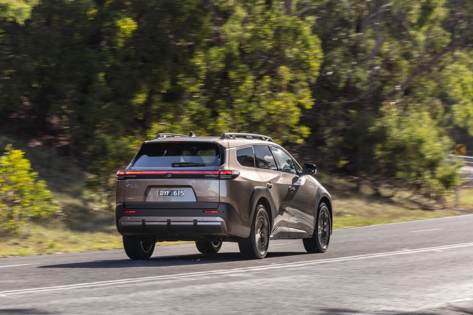 A bronze 2026 Toyota bZ4X PHEV SUV seen from the rear three-quarter angle while driving on a road surrounded by Australian bush
