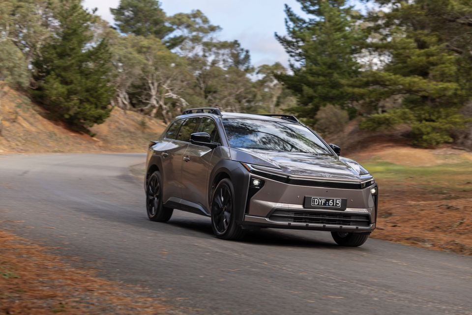 A grey 2026 Toyota bZ4X PHEV SUV driving on a winding road surrounded by trees, seen from the front
