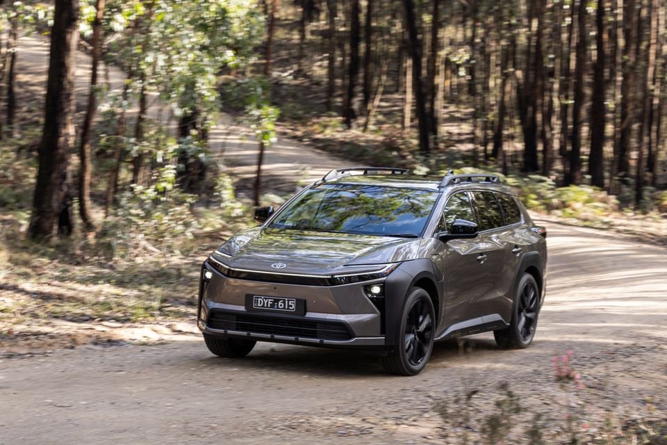 A grey 2026 Toyota bZ4X PHEV SUV driving along a tree-lined gravel road, seen from the front-left