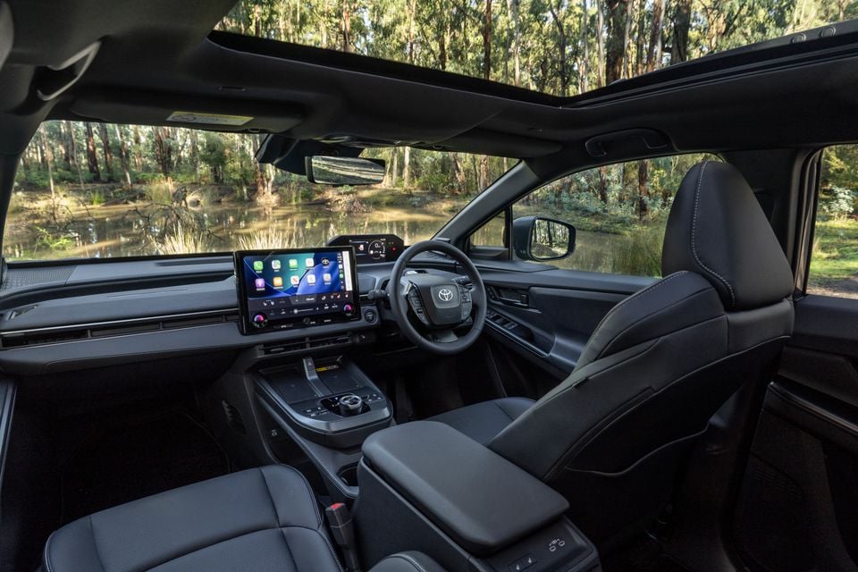 Interior of a Toyota bZ4X PHEV showing the dashboard, touchscreen infotainment system with Apple CarPlay, steering wheel, and dark upholstery, with a bushland water view through the windscreen