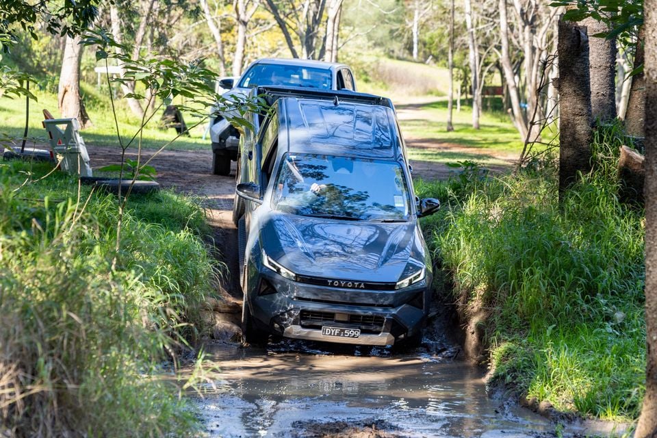 A dark-coloured Toyota Hilux navigating a muddy off-road track through bushland, seen from the front