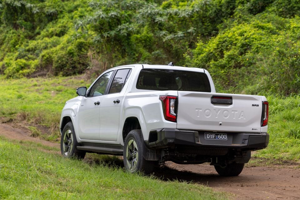 Um Toyota HiLux SR5 BEV de cabine dupla branco visto da traseira em um ângulo de três quartos em uma pista off-road de terra, cercada por uma vegetação verdejante.