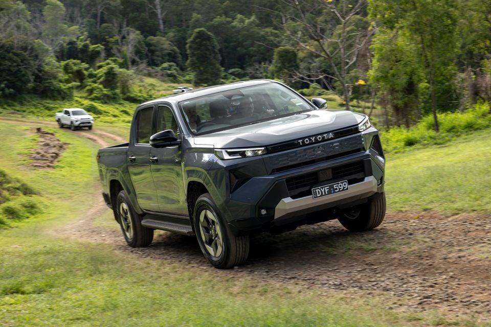 A grey 2026 Toyota HiLux BEV dual-cab ute driving along a dirt track surrounded by green bushland, seen from the front-side