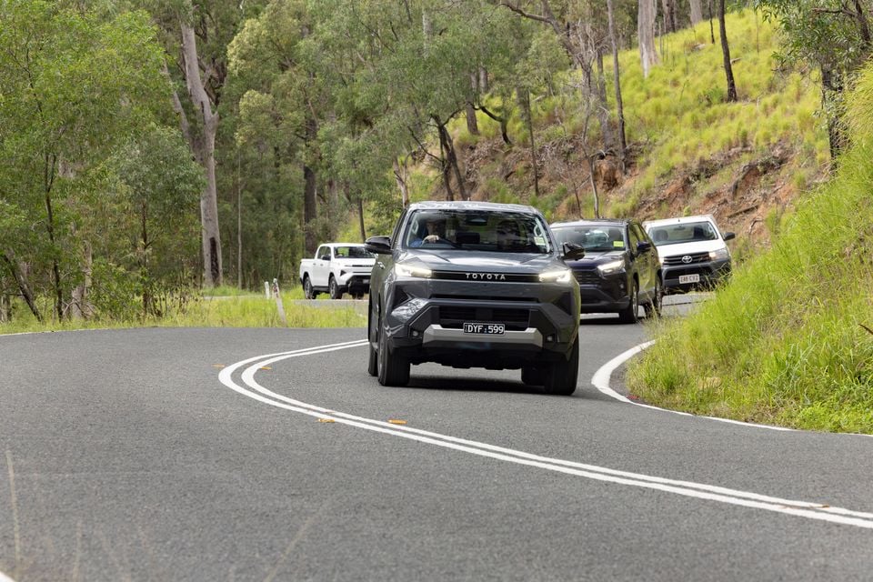 A dark grey Toyota Hilux ute driving along a winding rural road surrounded by Australian bush, seen from the front with other vehicles following behind