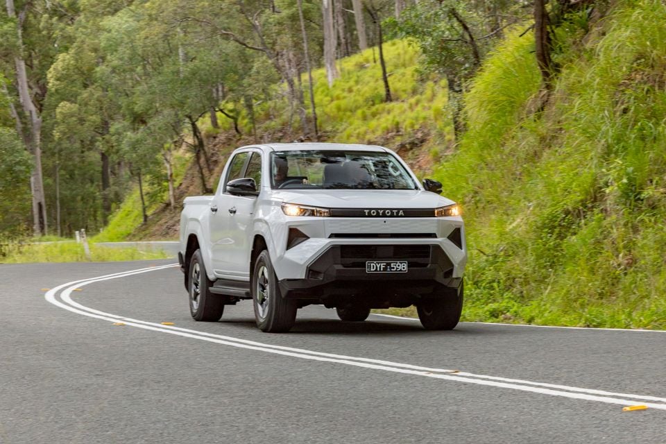 A white Toyota HiLux dual-cab ute driving along a winding bush road, seen from the front