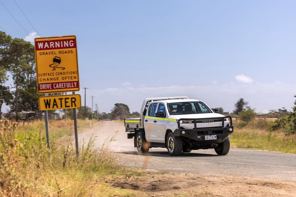 A white Toyota HiLux dual-cab ute with a flat tray and bull bar driving on a rural gravel road past an Australian warning sign