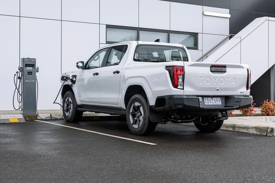 A white Toyota HiLux BEV dual-cab ute seen from the rear three-quarter angle, plugged into an Ocular DC fast charger in a car park