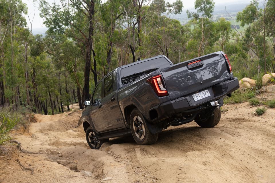 A dark grey Toyota HiLux BEV SR5 dual-cab ute seen from the rear, navigating a rough off-road dirt track surrounded by eucalyptus trees in Australia