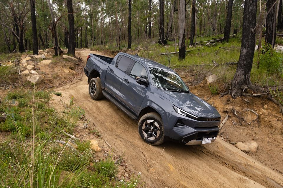 A grey 2026 Toyota HiLux BEV SR5 dual-cab ute navigating a dirt off-road track through an Australian bush setting, seen from the front-side