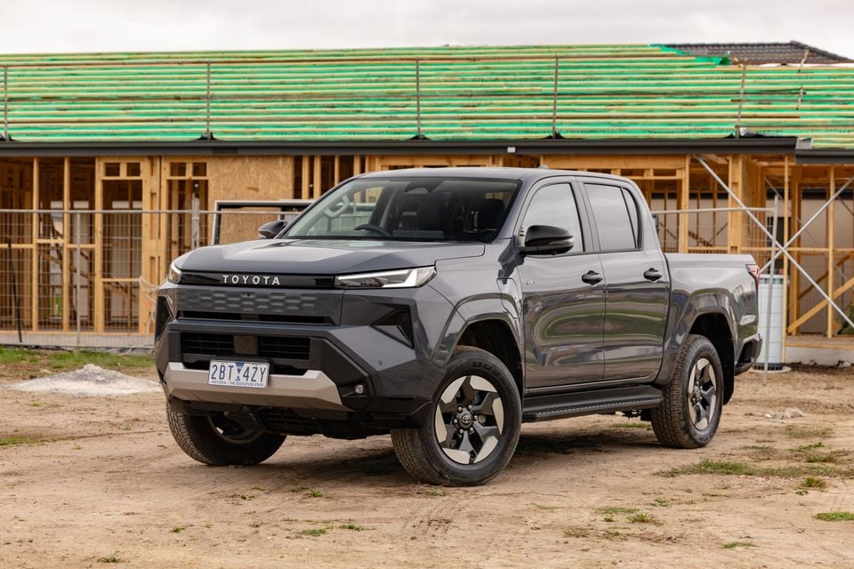 A grey 2026 Toyota HiLux BEV SR5 dual-cab ute parked on a dirt surface in front of a building under construction, seen from the front-side