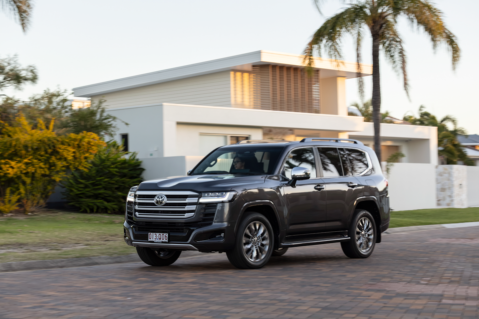 A dark grey Toyota LandCruiser 300 Series large SUV seen from the front-side angle, parked on a brick driveway in front of a modern home