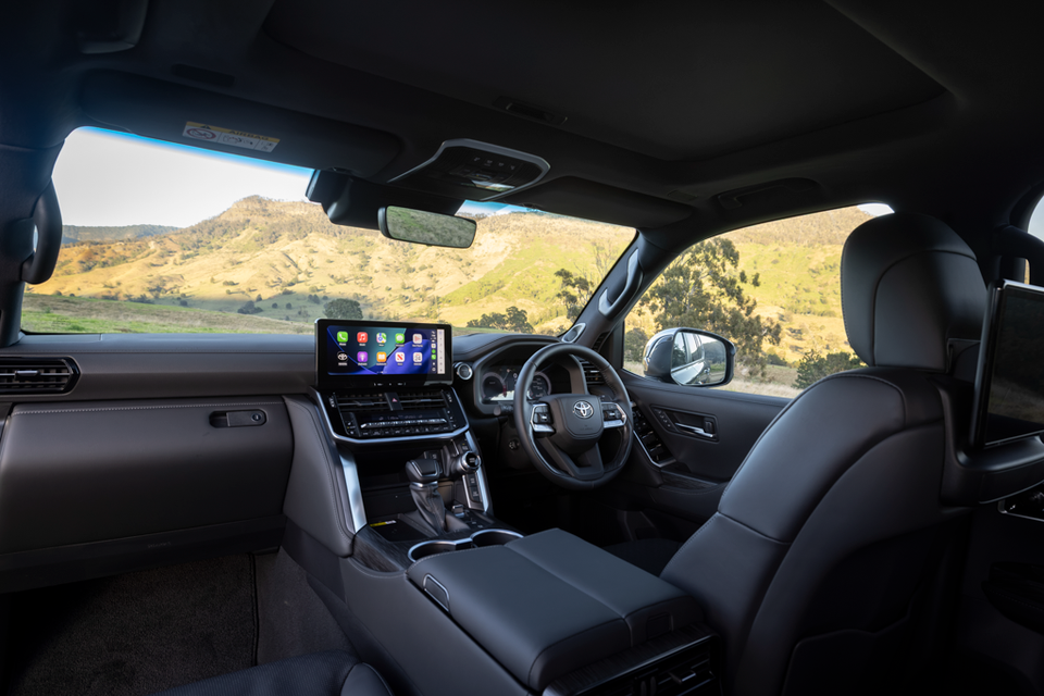 Interior of a Toyota LandCruiser 300 Series showing the dashboard, steering wheel, centre console, and touchscreen infotainment system displaying Apple CarPlay, with a scenic hilly landscape visible through the windows.