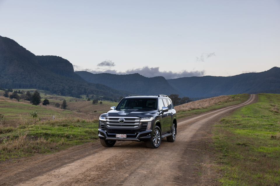 A dark grey Toyota LandCruiser 300 Series SUV seen from the front on a dirt road surrounded by green hills and mountains in rural Australia