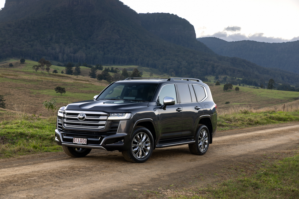 A dark grey Toyota LandCruiser 300 Series large SUV parked on a dirt road with green hills and mountains in the background