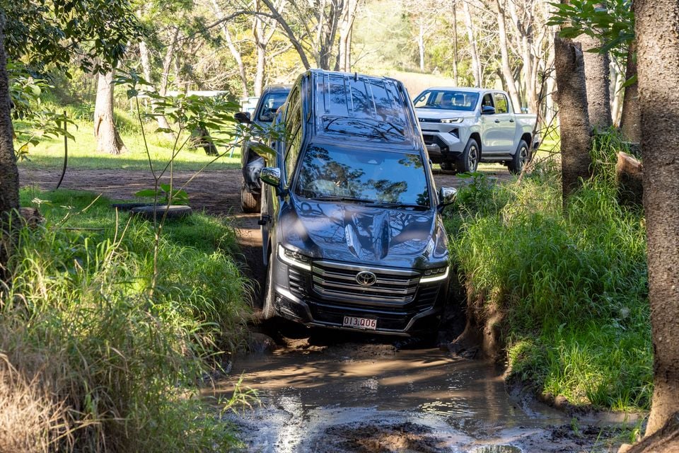 A dark-coloured 2026 Toyota LandCruiser SUV navigating a muddy off-road track through bushland, with a white ute visible in the background