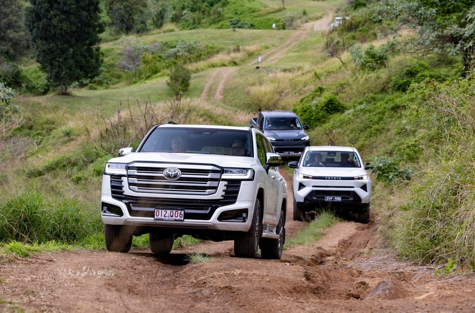 A white Toyota LandCruiser leads a convoy of SUVs along a rough dirt track through green hilly terrain during an off-road drive
