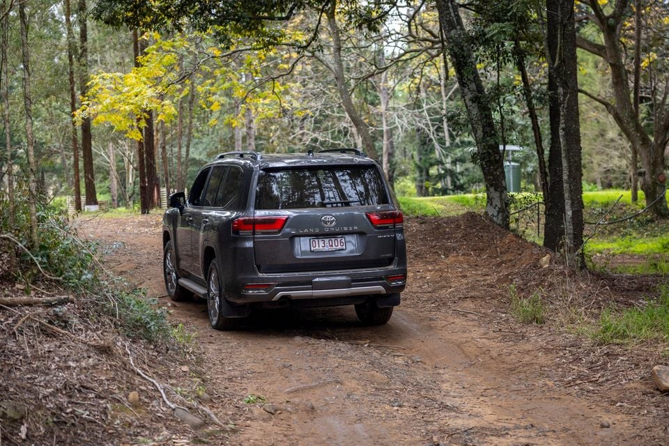 A grey Toyota Land Cruiser SUV seen from the rear, driving along a dirt track through a forest
