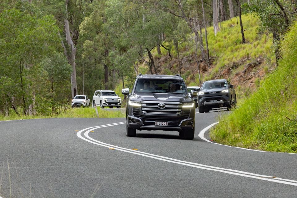 A dark grey Toyota LandCruiser SUV leads a convoy of Toyota vehicles along a winding road through Australian bushland
