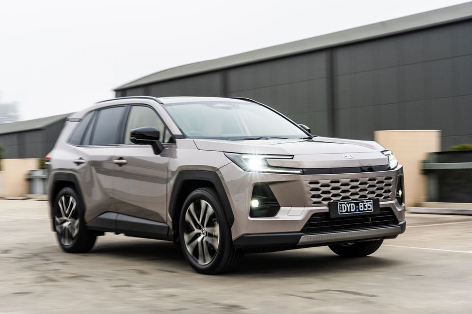 A beige 2026 Toyota RAV4 Cruiser SUV photographed from the front quarter while driving past an industrial building