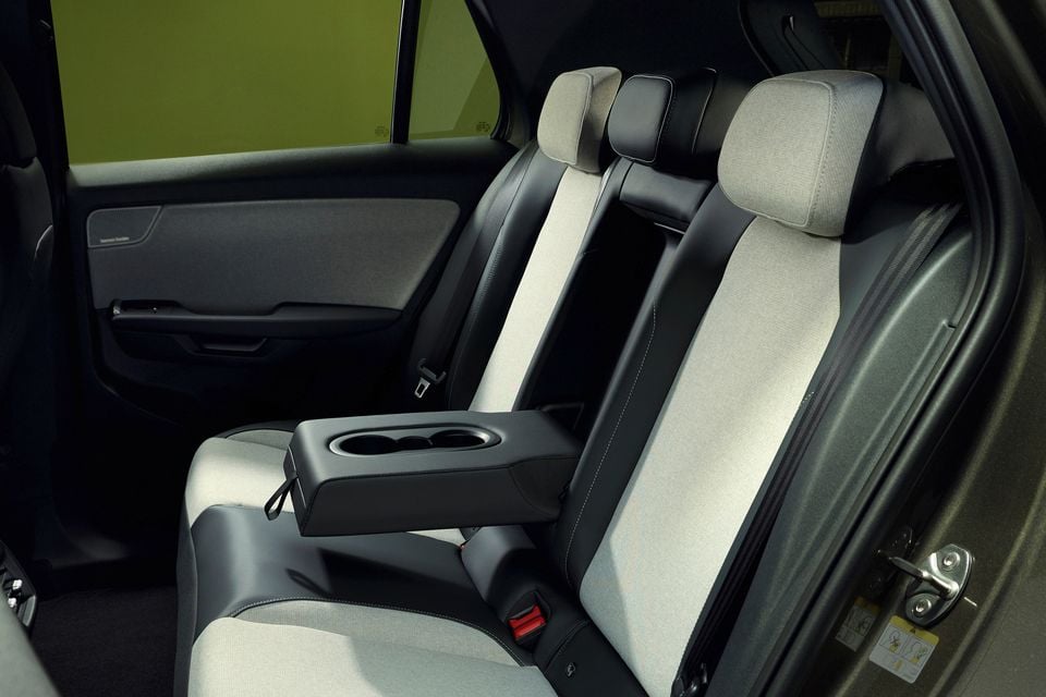 Interior do banco traseiro do Volkswagen ID 2026. Cruz, com estofamento em tecido cinza claro com detalhes em preto, apoio de braço central rebatível com porta-copos e painel da porta traseira.