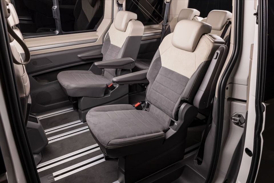 Interior of a 2026 Volkswagen Multivan showing rear passenger seats with two-tone grey and cream upholstery, armrests, and sliding door entry