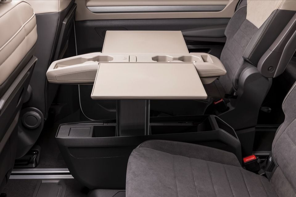 Rear interior of a 2026 Volkswagen Multivan showing a fold-out centre table with cup holders and under-floor storage between the second-row seats