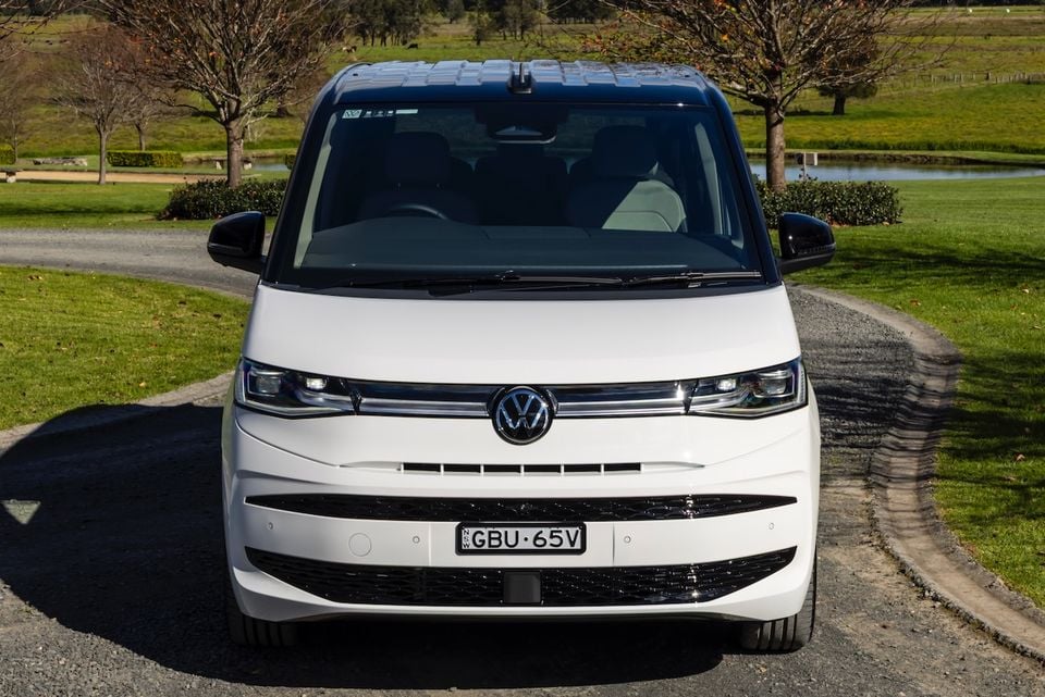 A white 2026 Volkswagen Multivan van seen from the front, parked on a gravel driveway surrounded by green parkland