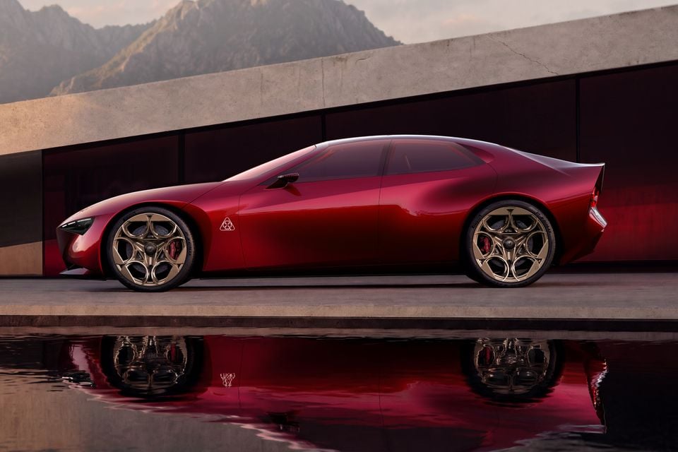 A red 2026 Alfa Romeo Cuorerosso coupe with bronze wheels seen in profile, reflected in a pool of water beside a concrete building
