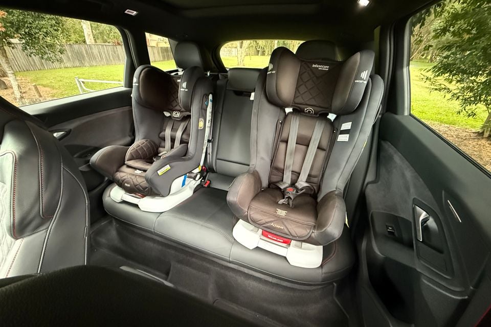 Two child car seats installed side by side on the rear seat of a 2026 Audi SQ6 e-tron, viewed from the open door with a garden visible through the windows