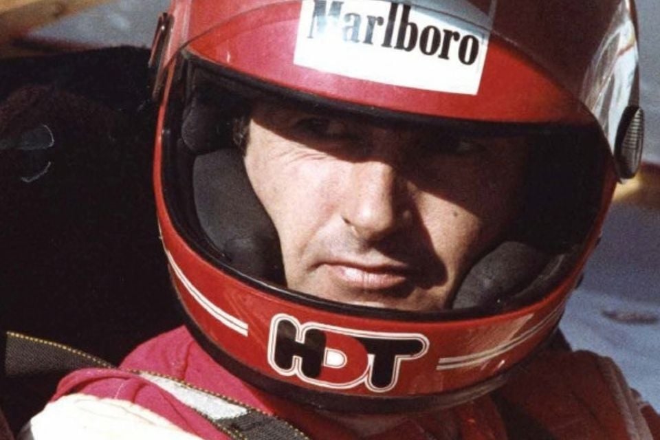 A racing driver wearing a red helmet with an HDT logo and Marlboro sticker sits in a race car cockpit.