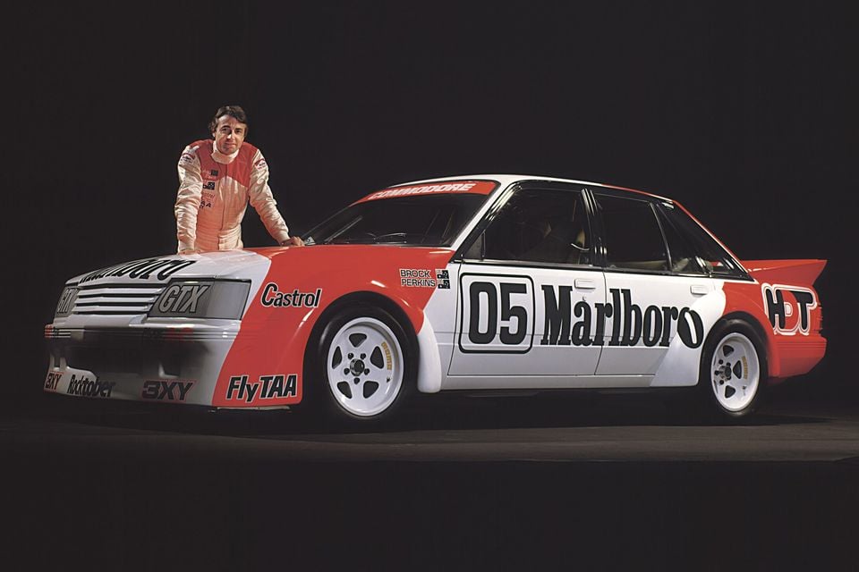 A driver stands beside a white and red 1980s Holden VK Commodore Group A touring car with Marlboro and HDT racing livery, photographed from the front quarter.