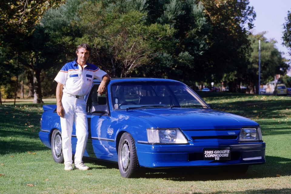 A man in white racing overalls stands next to a blue 1985 Holden Commodore Group A V8 parked on grass, seen from the front quarter angle.
