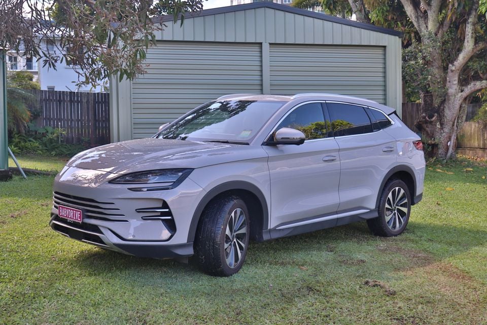 A silver 2026 BYD Sealion 6 SUV parked on grass in front of a green shed, seen from the front left side