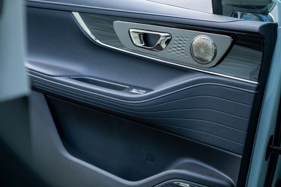 Close-up of a car door panel interior showing dark blue leather upholstery with wave stitching, a chrome door handle, wood trim, and a premium speaker grille