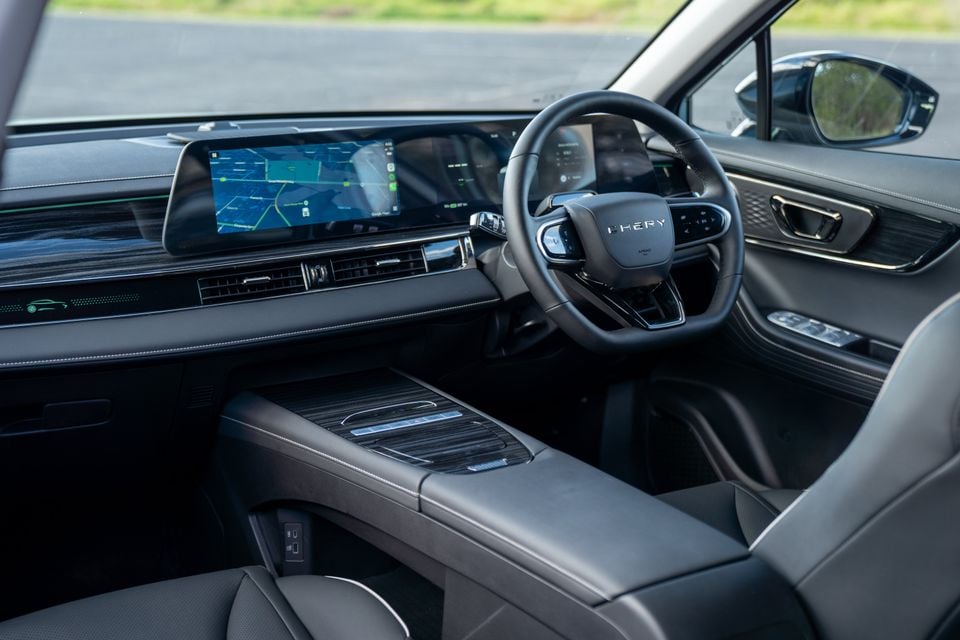 Interior of a Chery E5 showing the dashboard, wide dual touchscreen display with navigation, steering wheel with Chery branding, and dark leather upholstery