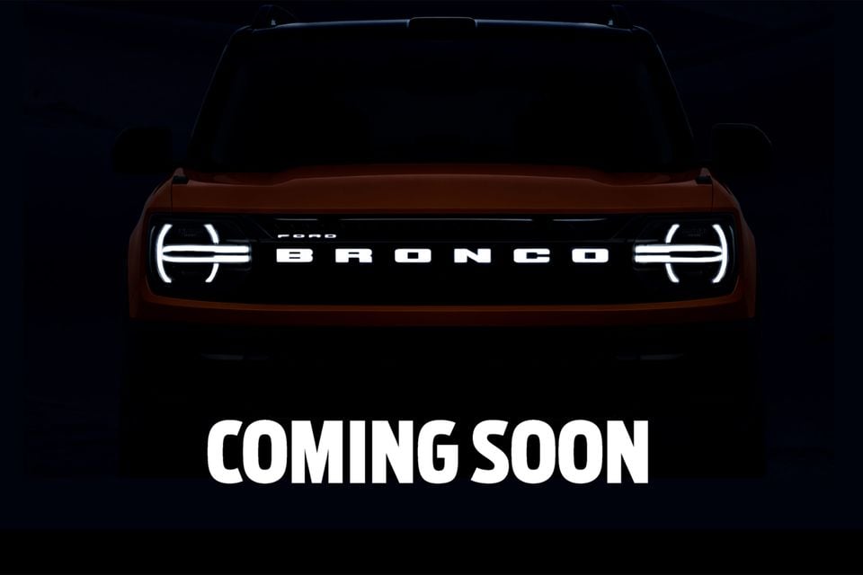 A teaser image of the Ford Bronco SUV seen from the front, partially silhouetted in darkness with illuminated headlights and 'BRONCO' lettering on the grille, with 'Coming Soon' text below