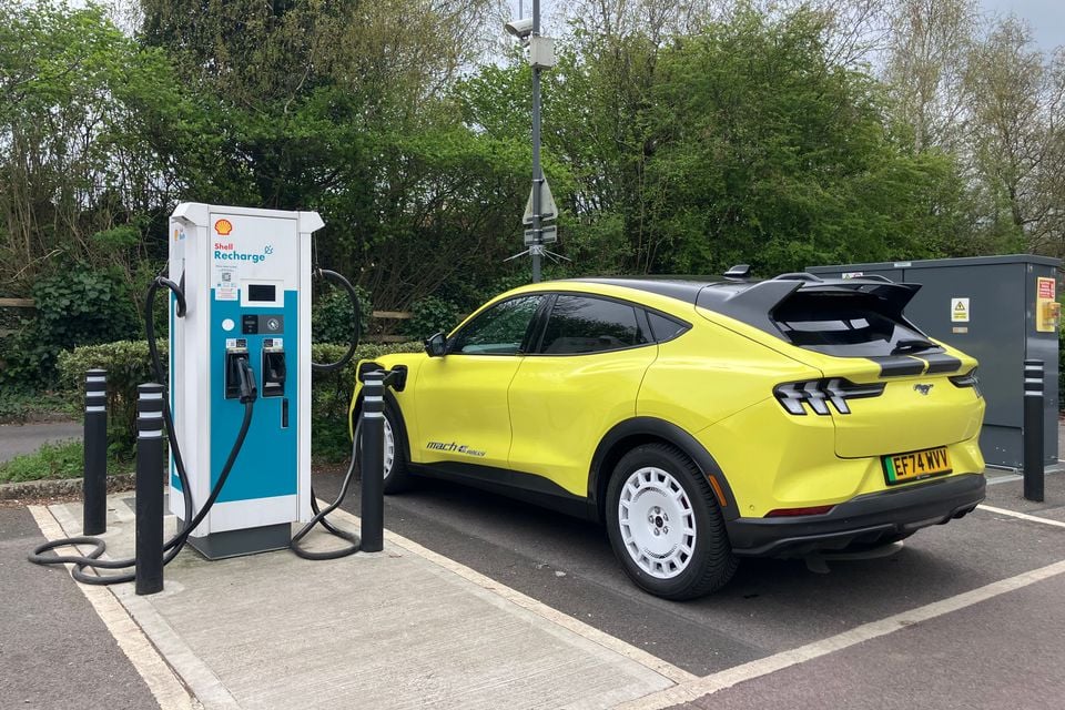A yellow Ford Mustang Mach-E Rally SUV charging at a Shell Recharge public electric vehicle charging station in a car park, seen from the rear three-quarter angle