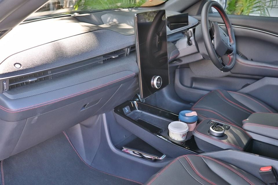 Interior of a Ford Mustang Mach-E showing the centre console, large vertical touchscreen, steering wheel with red stitching, and cup holders with drinks in them