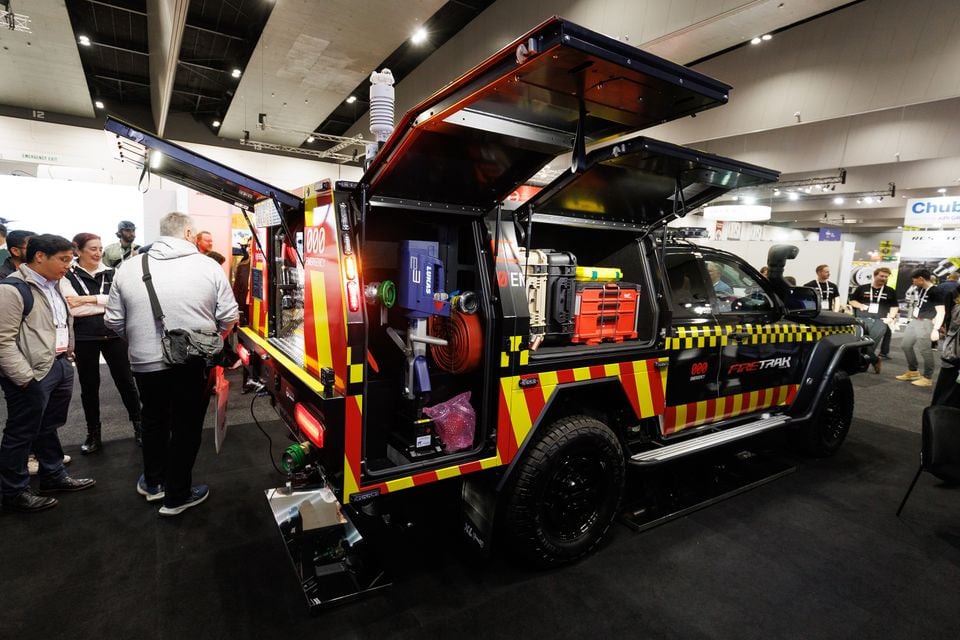 A Ford Ranger Super Duty fire response unit with red, yellow and black emergency livery and open storage compartments loaded with firefighting equipment, on display at an indoor expo with attendees looking on
