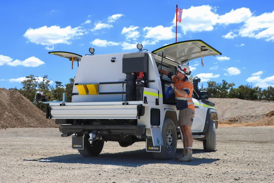 A worker in hi-vis clothing and hard hat accessing the toolbox fitted to a white ute with a custom tray and canopy setup at a mining or construction site